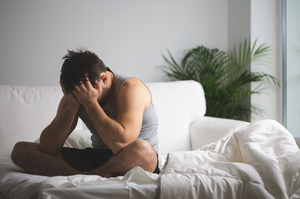 Homem jovem sentado na beira da cama, com a cabeça entre as mãos, em um gesto que representa uma crise de ansiedade